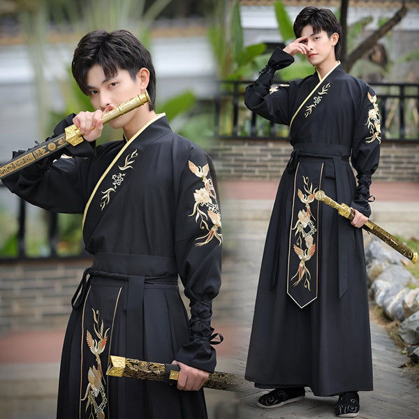 Un jeune homme en costume traditionnel chinois noir avec broderies dorées, tenant un fourreau d'épée doré près de sa bouche et posant avec une épée, devant un arrière-plan flou de verdure et d'architecture traditionnelle.