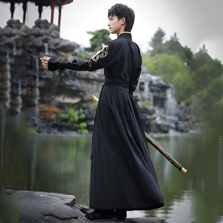 Un homme en costume traditionnel noir avec une épée, debout sur une pierre au bord d'un lac. Le costume a des broderies dorées sur la manche. Un paysage asiatique avec des arbres et une cascade est visible en arrière-plan.