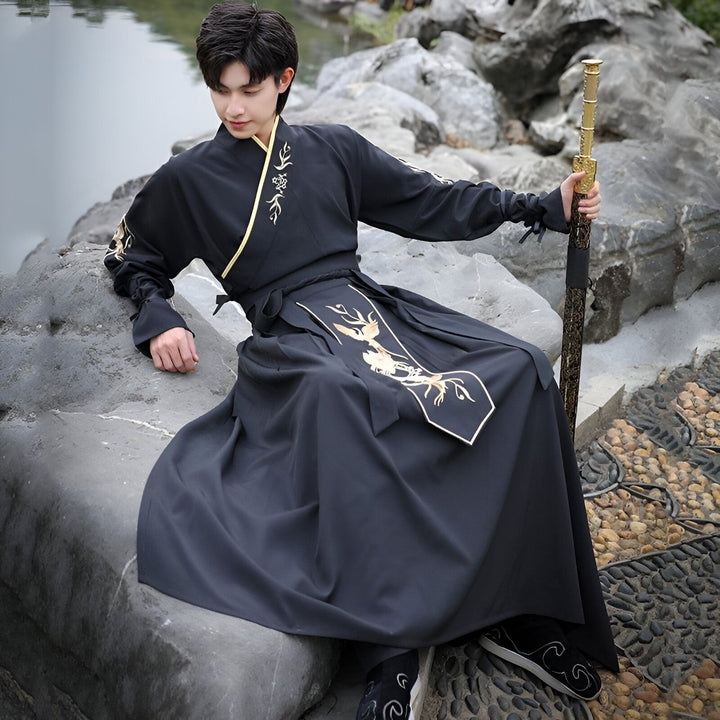 Un homme en costume traditionnel asiatique noir et or, assis sur un rocher avec une épée. Il a un regard calme et son costume est richement décoré de motifs dorés. L'arrière-plan est flou, avec de la végétation et de l'eau.
