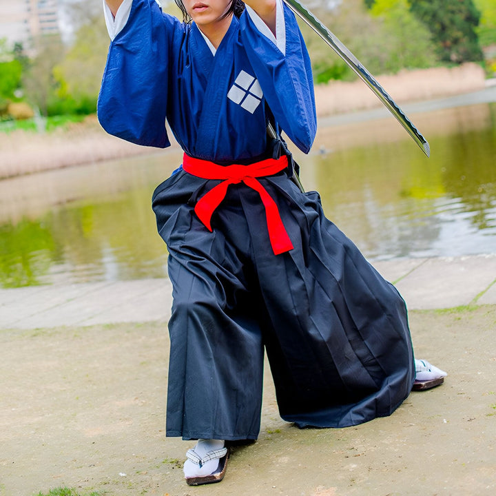 Homme en kimono bleu traditionnel avec ceinture rouge et hakama noir, tenant un katana. Idéal pour cosplay ou arts martiaux. Découvrez notre collection de kimonos authentiques.