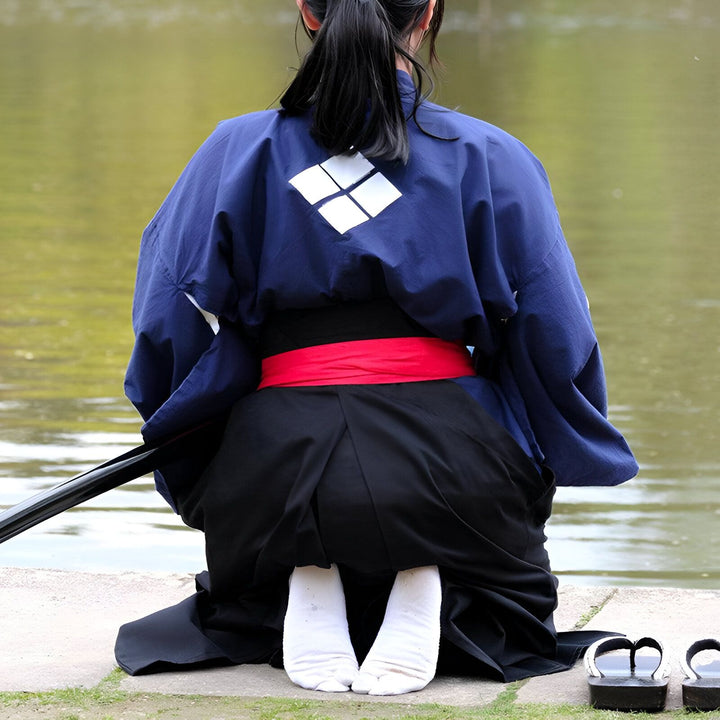 Femme à genoux en kimono bleu avec motif losange blanc, ceinture rouge et hakama noire. Sandales geta à côté. Vêtements traditionnels japonais.