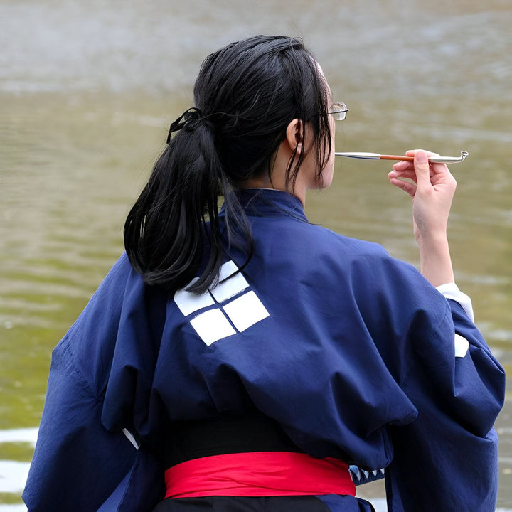 Femme portant un kimono bleu traditionnel avec motif blanc et ceinture rouge, fumant une pipe. Découvrez notre collection de kimonos authentiques.