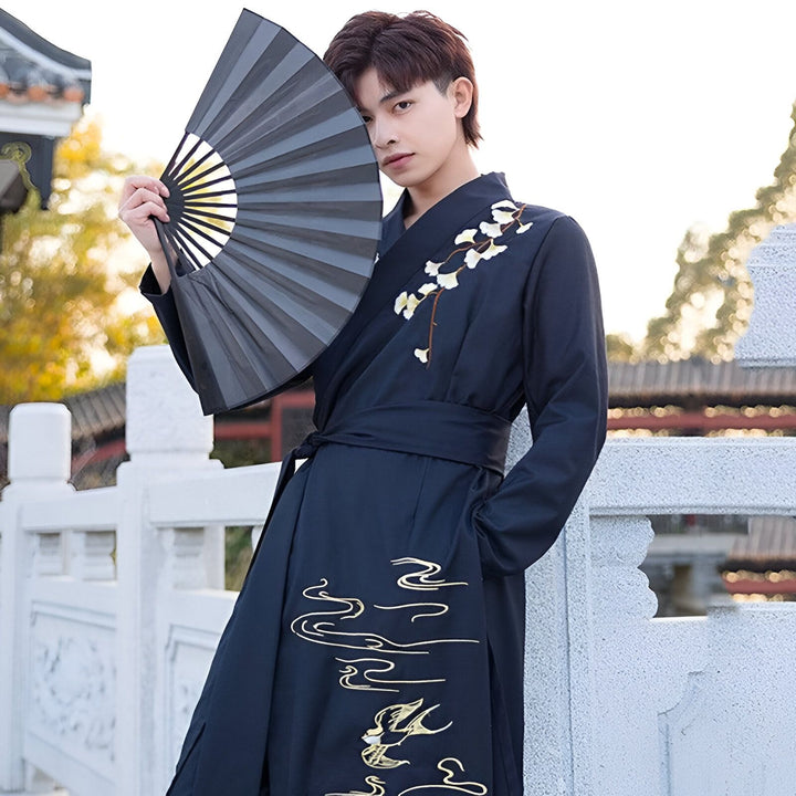 Homme portant un kimono bleu marine brodé avec des motifs floraux et d'oiseaux, tenant un éventail noir. Kimonos traditionnels et modernes disponibles à l'achat.