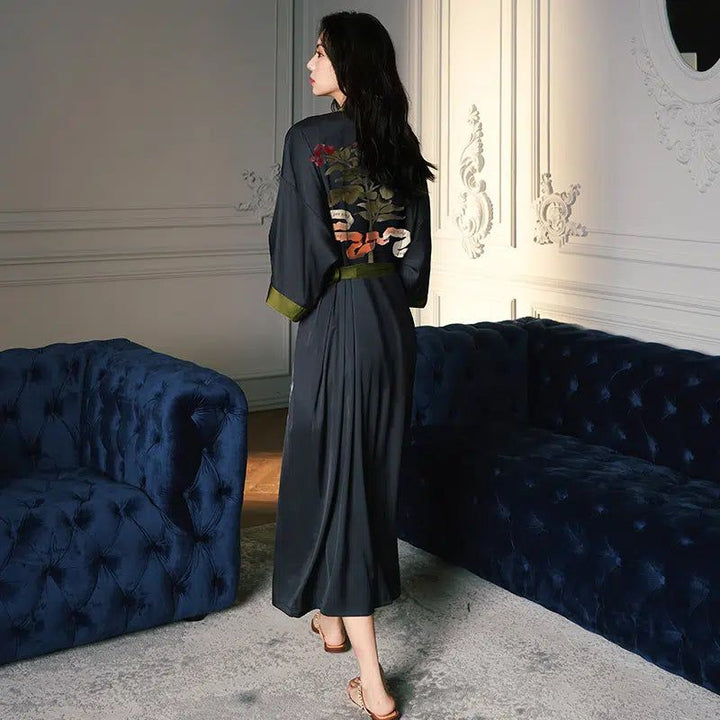 Femme portant un kimono gris foncé avec des motifs floraux brodés dans le dos. Élégant peignoir long, idéal pour la détente ou comme vêtement d'intérieur raffiné.
