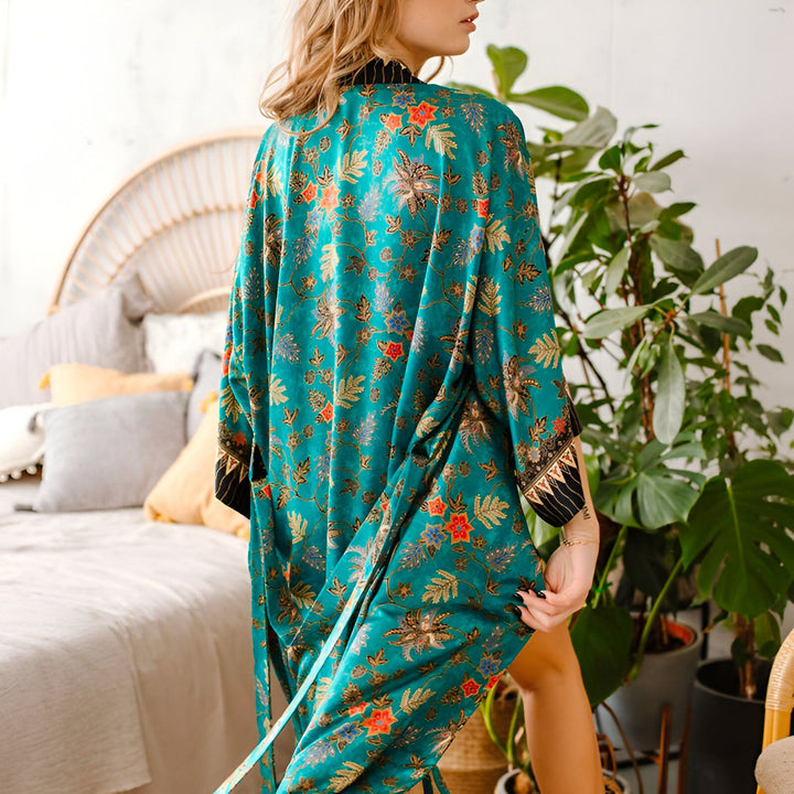 Femme portant un kimono turquoise à motifs floraux. Vue de dos, ambiance intérieure avec lit et plantes. Kimono élégant et confortable pour la maison. Découvrez notre collection de kimonos uniques.