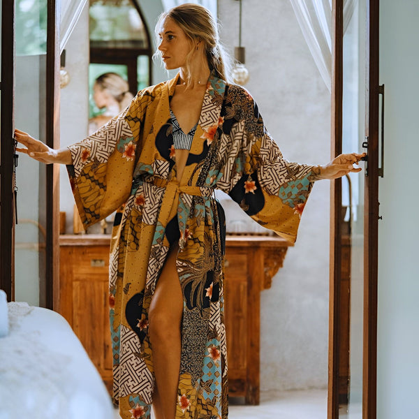 Femme blonde ouvrant une porte, portant un kimono long à motifs jaunes, noirs, bleus et rouges. Kimono de notre collection disponible à l'achat.