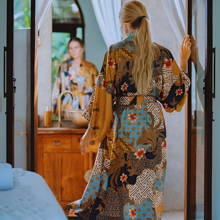Femme portant un kimono à motifs colorés, vue de dos. Le kimono est bleu, or et noir avec des motifs floraux. Une autre femme, portant un kimono similaire, se reflète dans un miroir. Kimonos élégants pour toutes occasions.