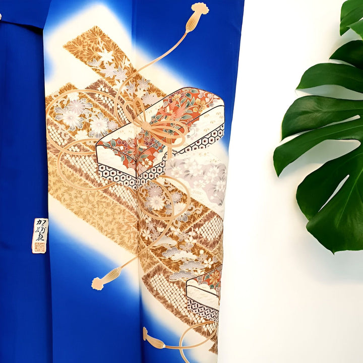 Détail d'un kimono bleu avec motifs de boîtes décoratives et fleuries, nouées par un cordon doré. Feuille de monstera en arrière-plan. Kimonos traditionnels et modernes en vente.