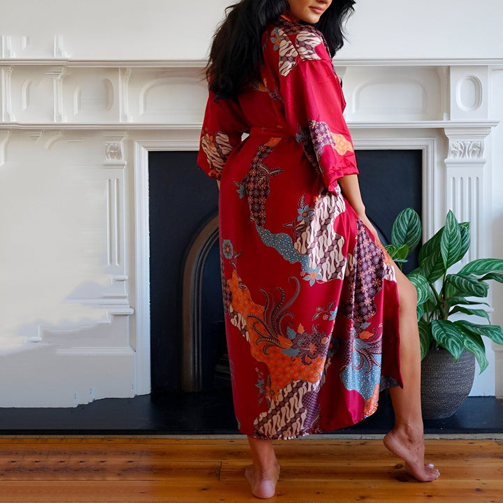 Femme portant un kimono rouge à motifs devant une cheminée. Kimono long, ample, parfait pour la maison ou comme peignoir. Découvrez notre collection de kimonos!