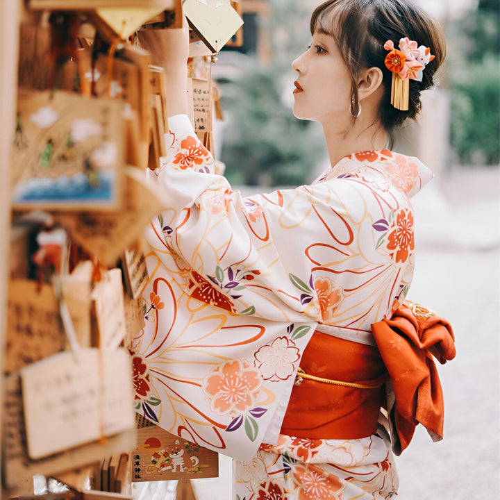 Femme asiatique en kimono traditionnel blanc à motifs floraux, avec obi orange. Cheveux ornés d'une fleur. Elle examine des plaques votives en bois. Kimonos japonais authentiques en vente.