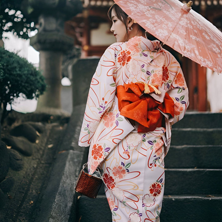 Femme portant un kimono traditionnel japonais à motifs floraux avec obi rouge. Elle tient un parapluie décoratif et monte des escaliers. Kimonos de haute qualité disponibles à l'achat.