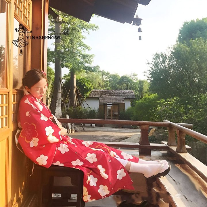 Femme en kimono rouge à motifs floraux blancs, assise sur un balcon avec vue sur jardin japonais. Kimono traditionnel japonais à vendre. Vêtements japonais authentiques.