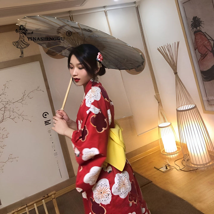 Femme portant un kimono rouge à motifs floraux blancs et marrons, avec un obi jaune. Elle tient un parasol japonais. Kimonos traditionnels à vendre.
