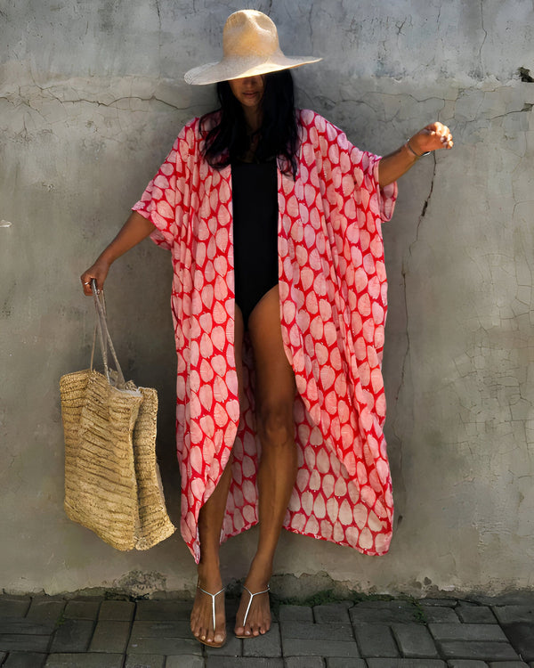 Femme portant un kimono rouge à motifs de feuilles, chapeau de paille, et maillot de bain noir. Sac de plage en paille. Vente de kimonos de plage tendance et confortables.