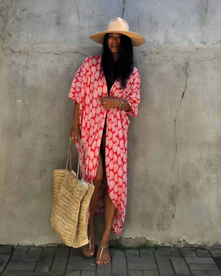 Femme souriante portant un kimono long rouge et blanc à motifs, un chapeau de paille et un sac en paille. Look estival décontracté. Vente de kimonos en ligne.