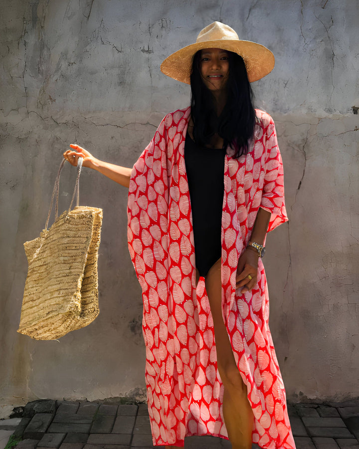 Femme portant un kimono rouge imprimé, un maillot de bain noir et un chapeau de paille. Elle tient un sac de plage en paille. Kimonos de qualité à découvrir sur notre site.