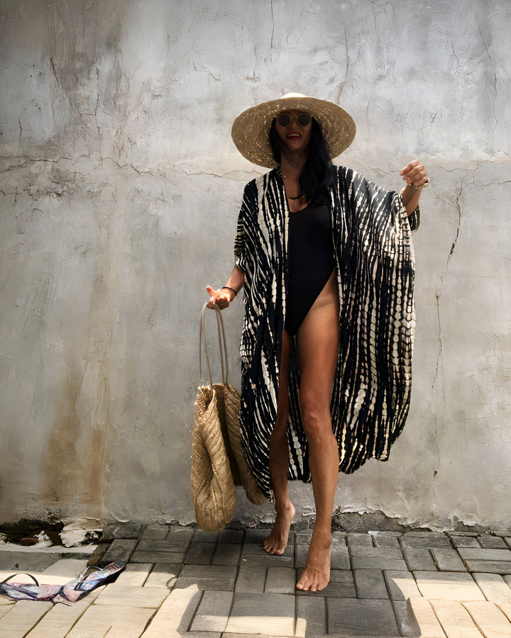 Femme portant un kimono noir et blanc avec un maillot de bain noir, un chapeau de paille et des lunettes de soleil. Elle tient un sac de paille. Mode kimono, vêtements de plage.