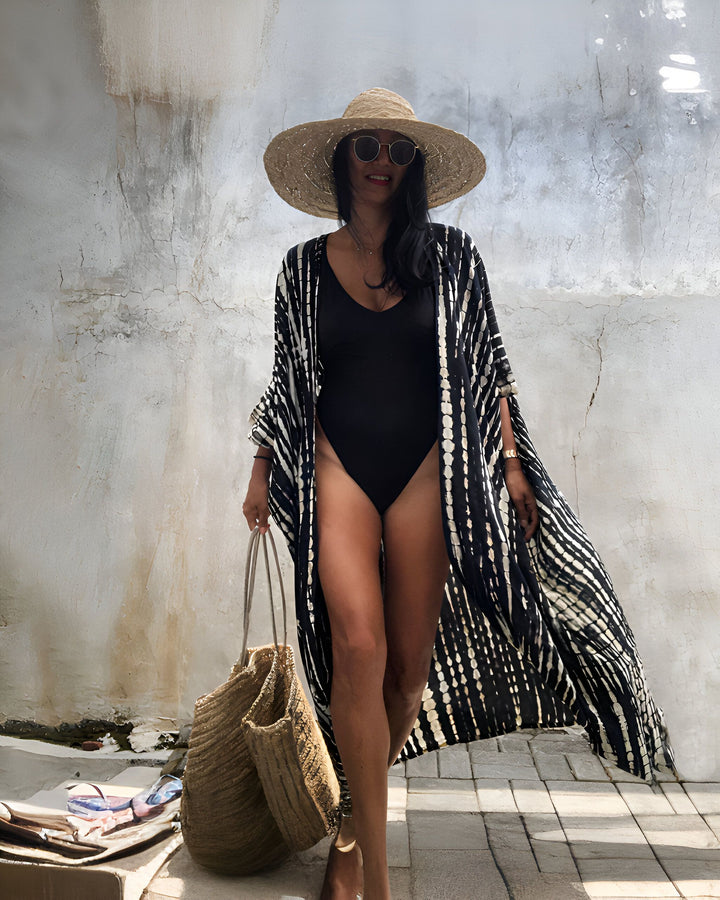 Femme portant un kimono noir et blanc à motifs, un maillot de bain noir, un chapeau de paille et des lunettes de soleil. Sac de plage en paille. Mode estivale, vêtements de plage. Kimonos en vente.