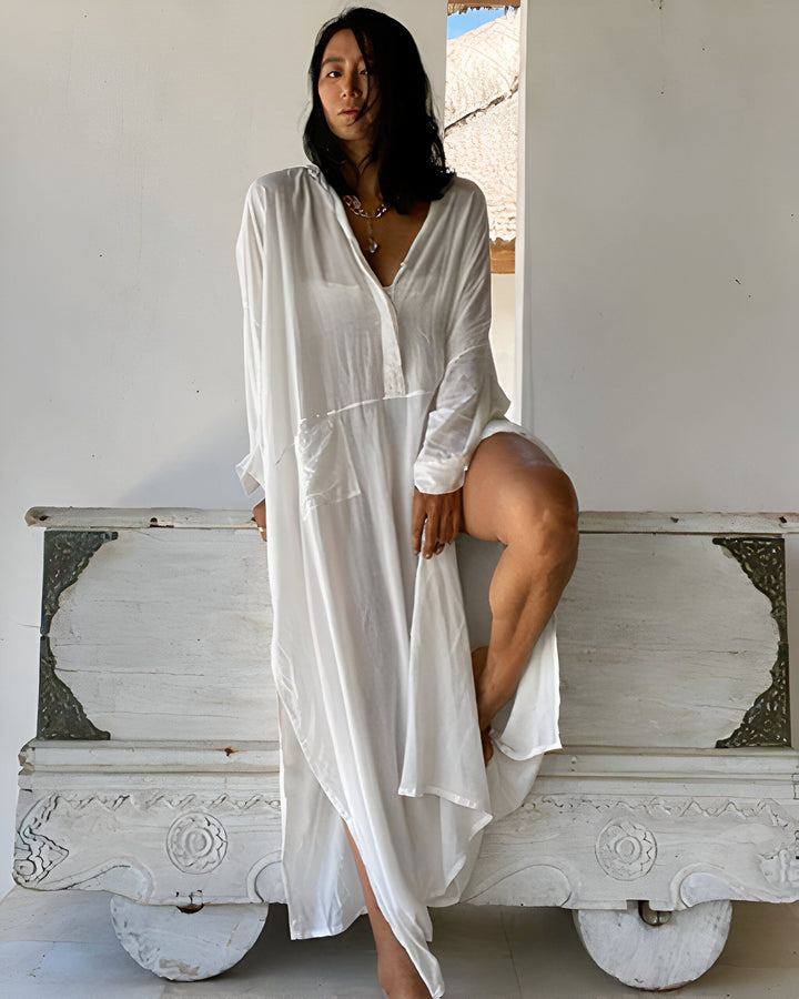 Femme portant un kimono blanc ample, assise sur un coffre en bois blanc. Collier argenté. Vêtements de détente, style kimono. Achetez nos kimonos confortables et élégants.