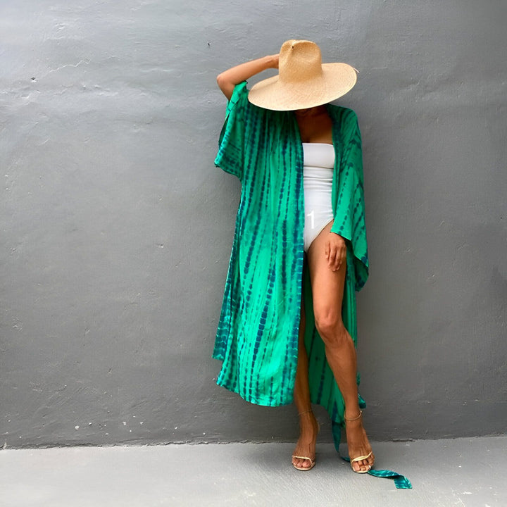 Femme en kimono vert et bleu tie-dye, maillot de bain blanc et chapeau de paille. Kimonos élégants pour la plage ou la maison. Découvrez notre collection de kimonos uniques.