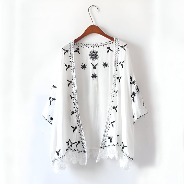 Kimono blanc brodé de motifs floraux noirs, suspendu à un cintre en bois. Détails en dentelle. Vêtement léger et élégant. Notre collection de kimonos.