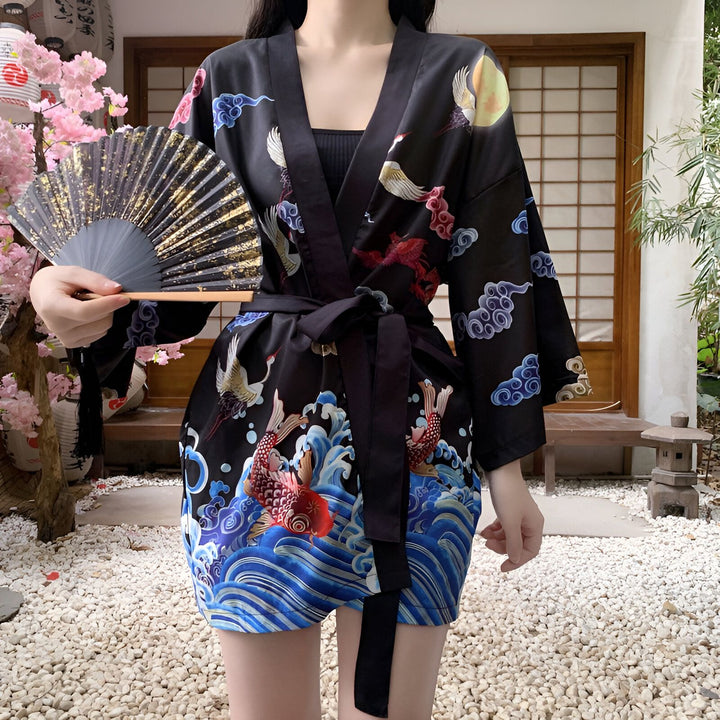 Femme portant un kimono noir avec motifs japonais traditionnels : carpes koï, grues, vagues et nuages. Elle tient un éventail doré. Vêtements japonais, boutique de kimonos.