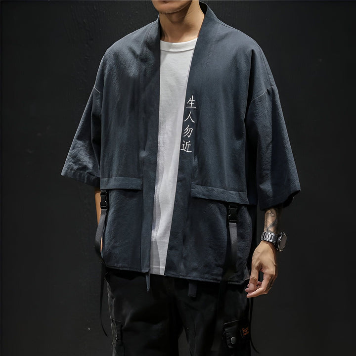 Homme portant un kimono gris anthracite avec inscription verticale blanche. Vêtement de style japonais avec poches et lanières. Collection de kimonos pour homme.