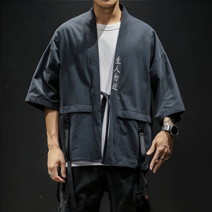 Homme portant un kimono gris anthracite avec des inscriptions blanches. Veste kimono style japonais avec poches et attaches, idéale pour un look mode et décontracté. Découvrez notre collection de kimonos pour hommes.