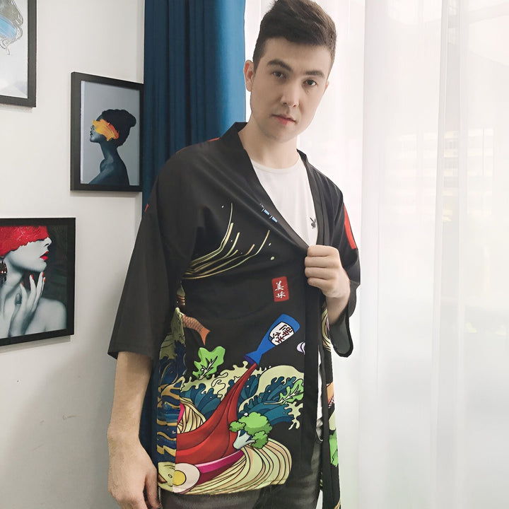 Homme portant un kimono noir imprimé de motifs ramen, devant un rideau et des tableaux. Kimonos pour hommes disponibles sur notre boutique en ligne.
