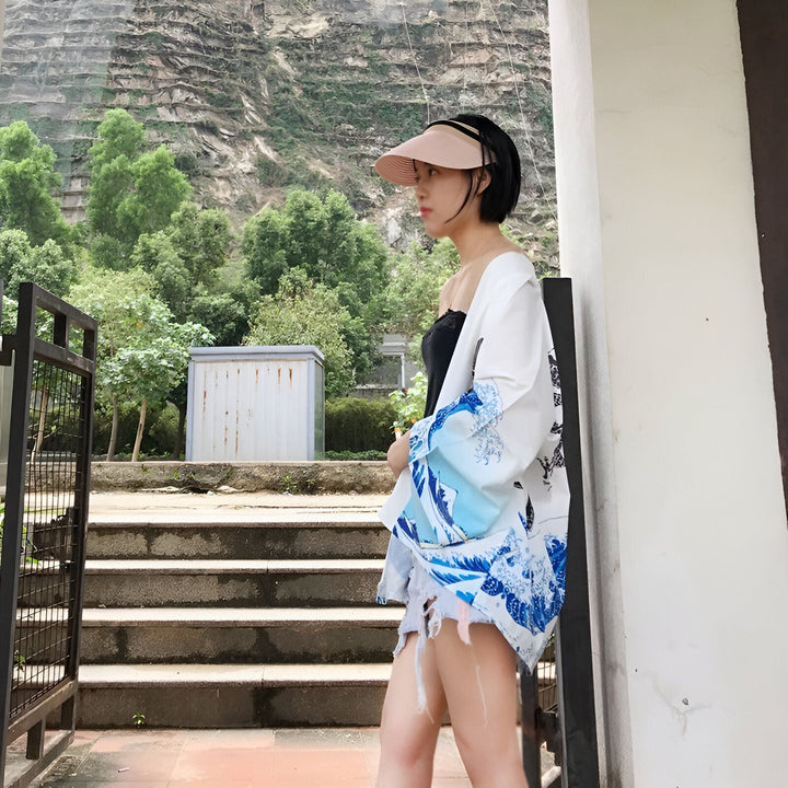 Femme portant un kimono blanc avec des motifs de vagues bleues, un débardeur noir et un chapeau de soleil, devant un paysage montagneux. Kimonos pour femmes disponibles à l'achat.