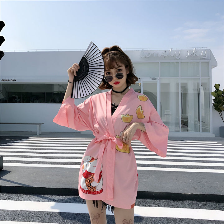 Femme portant un kimono rose avec motifs chat porte-bonheur et lingots d'or, tenant un éventail noir et blanc. Vêtements de style japonais disponibles à l'achat.