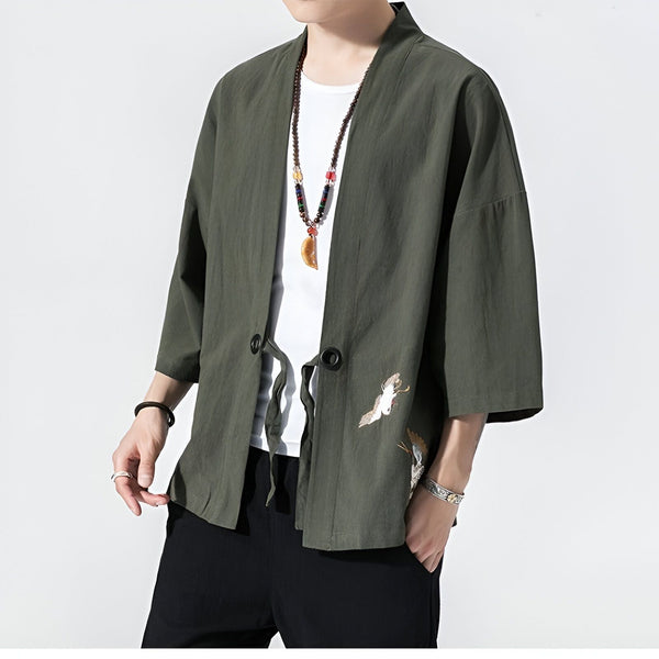 Homme portant un kimono vert olive avec des motifs de grues blanches brodés. Vêtement de style japonais avec cordon de fermeture et manches 3/4. Colliers et bijoux.