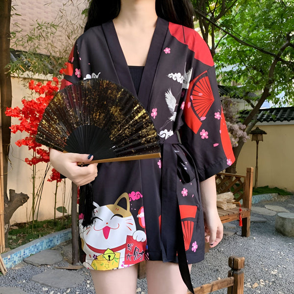 Kimono noir à motifs japonais (chats, grues, éventails) porté par une femme, avec un éventail doré à la main. Vêtements japonais.