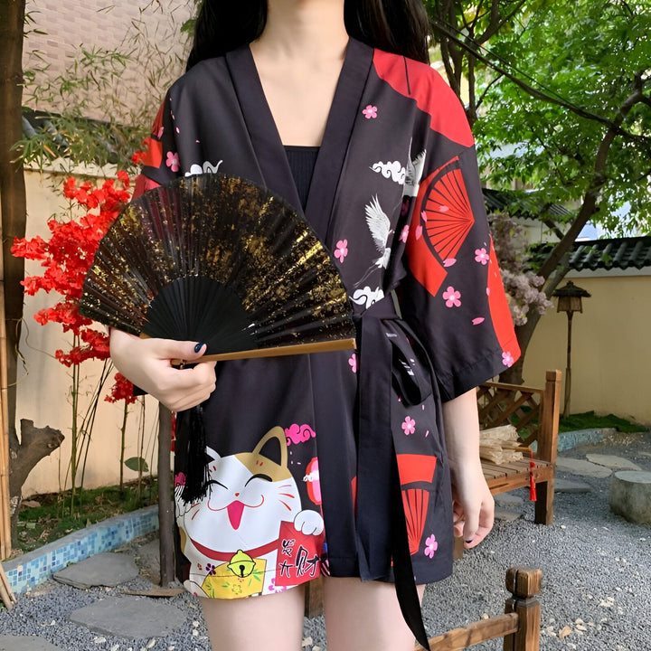 Kimono noir à motifs japonais (chats, grues, éventails) porté par une femme, avec un éventail doré à la main. Vêtements japonais.