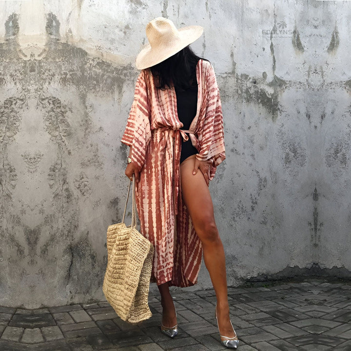 Femme portant un kimono long imprimé tie-dye, chapeau de paille et sac en paille. Mode kimono pour la plage ou une tenue décontractée. Boutique en ligne de kimonos tendance.