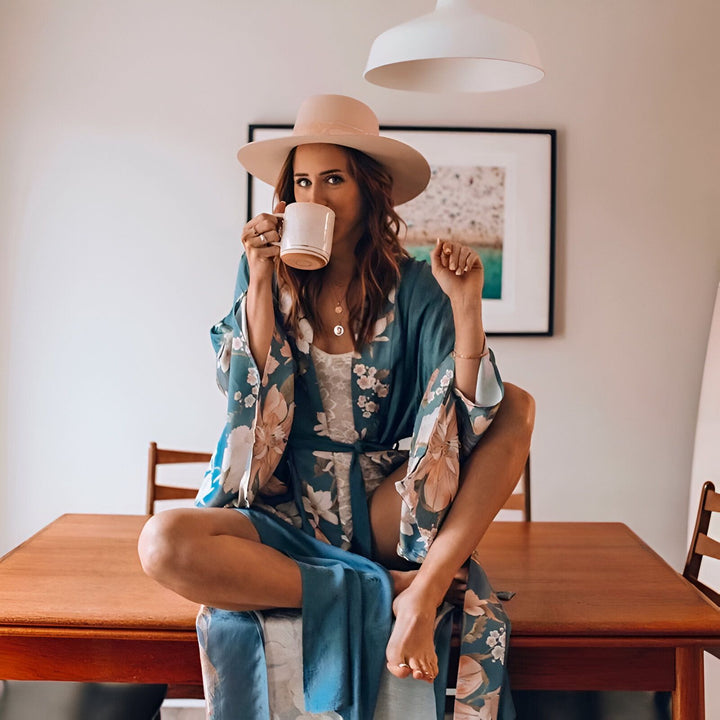 Femme assise sur une table en kimono fleuri bleu, buvant du café, avec un chapeau. Découvrez notre collection de kimonos uniques.