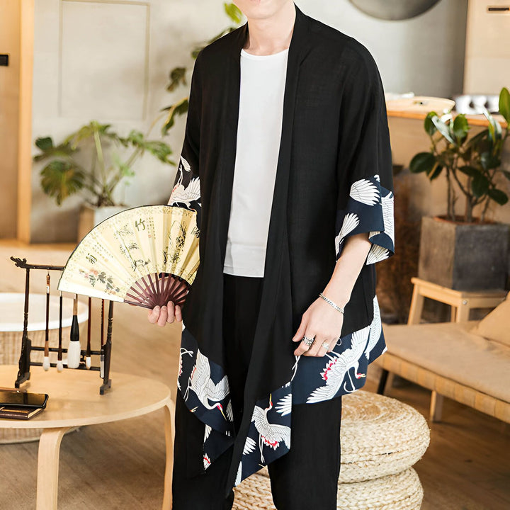 Homme portant un kimono noir avec des motifs de grues blanches et bleues, tenant un éventail. Mode kimono.