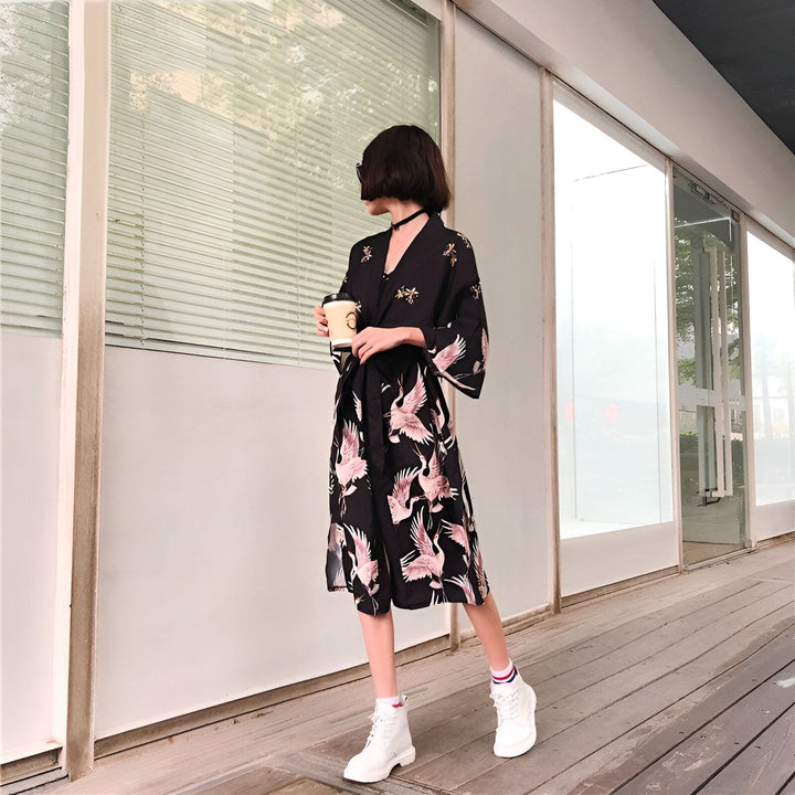 Femme portant un kimono noir à motifs de grues roses, tenant une tasse de café. Vêtements de style japonais à la mode. Kimonos pour femmes disponibles à l'achat.
