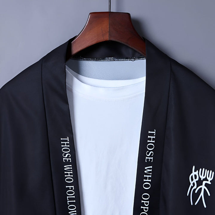 Kimono noir avec motifs blancs et lettrage, suspendu à un cintre. Vêtement de style japonais, idéal pour un look moderne et décontracté. Découvrez notre collection de kimonos uniques.