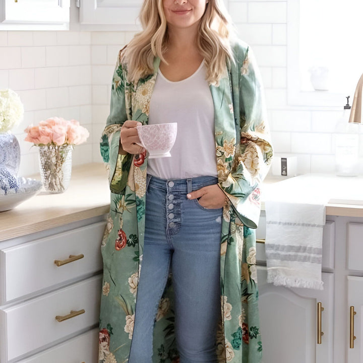 Femme portant un kimono fleuri vert et une tasse de thé dans une cuisine lumineuse. Notre collection de kimonos chics pour toutes les occasions.