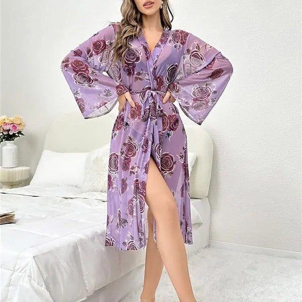 Femme portant un kimono lilas transparent à motifs floraux devant un lit. Kimono avec manches évasées et ceinture assortie. Vêtements de nuit, peignoir, produit kimono.