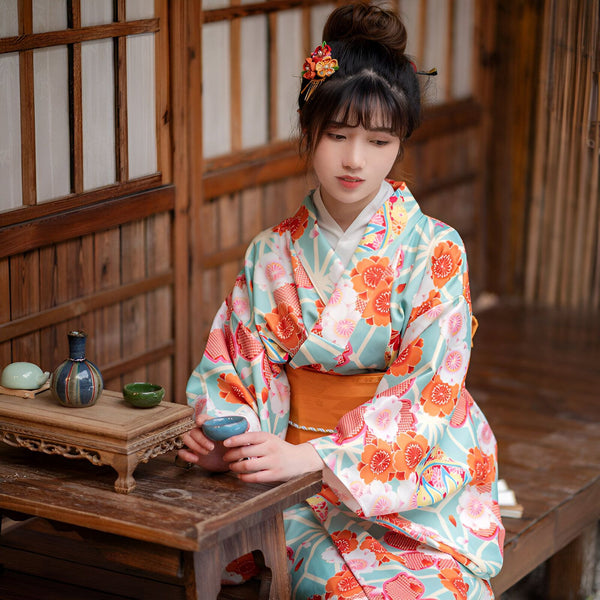 Femme asiatique portant un kimono floral coloré. Détails soignés : chignon orné, obi orange. Vente de kimonos authentiques.