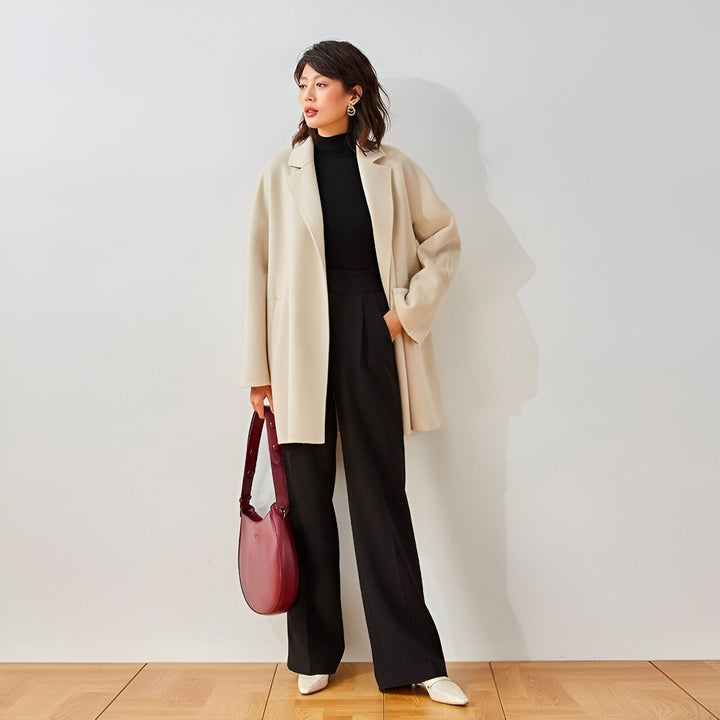 Femme portant un manteau beige, un col roulé noir et un pantalon noir ample. Elle tient un sac à main bordeaux. Style kimono moderne, élégant et confortable.