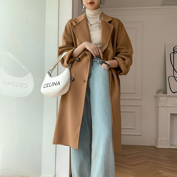 Femme portant un long manteau marron, un col roulé blanc et un jean. Sac à main blanc visible. Mode automne-hiver, style kimono moderne.