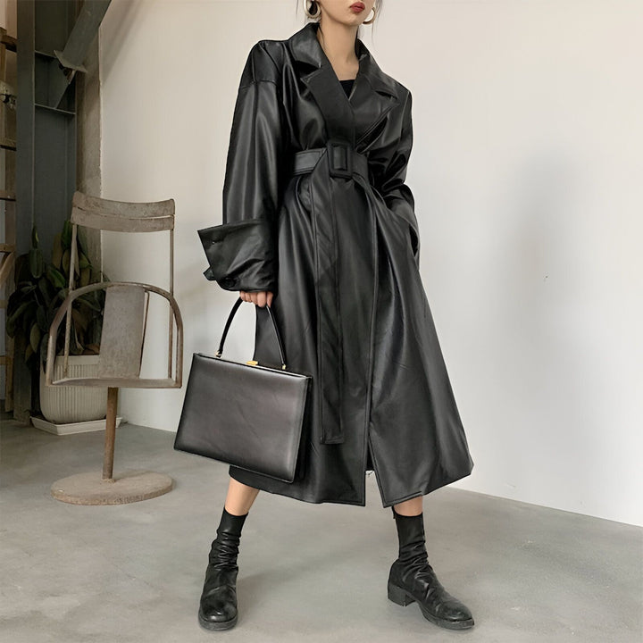 Femme portant un long kimono noir en cuir avec une ceinture et tenant un sac à main noir. Élégance et style kimono moderne.
