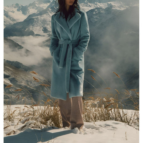 Femme portant un kimono long bleu ciel ceinturé, devant un paysage montagneux enneigé. Mode hivernale, vente de kimonos en ligne.