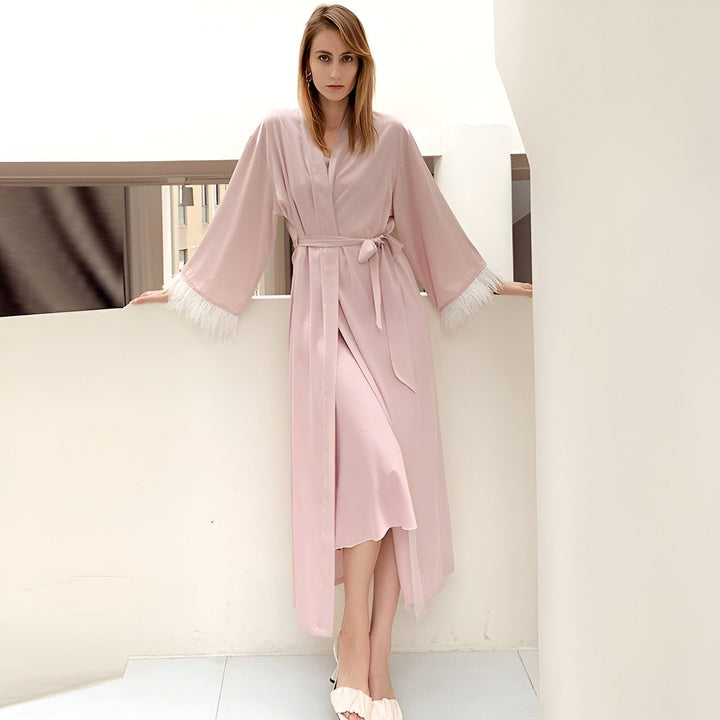 Femme portant un kimono rose poudré avec des plumes blanches aux manches. Élégant peignoir de notre collection. Kimonos de qualité à découvrir.