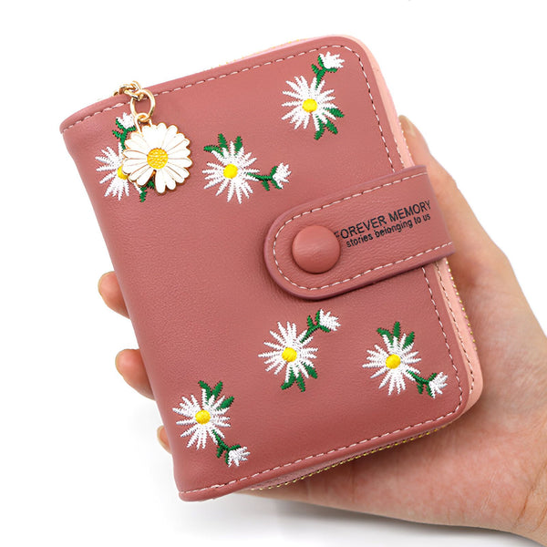 Gros plan sur un portefeuille rose orné de motifs floraux de marguerites blanches. Un charm en forme de marguerite pend du coin supérieur gauche.