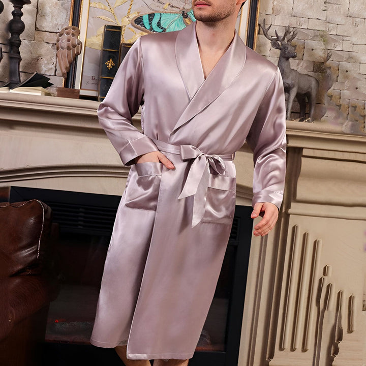 Homme portant un kimono long en satin rose pâle avec une ceinture nouée, devant une cheminée. Vente de kimonos de luxe en ligne.