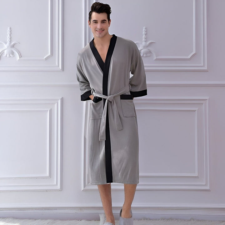 Homme souriant portant un kimono/peignoir gris avec garniture noire et ceinture assortie. Vêtements de détente confortables et élégants disponibles sur notre boutique en ligne.
